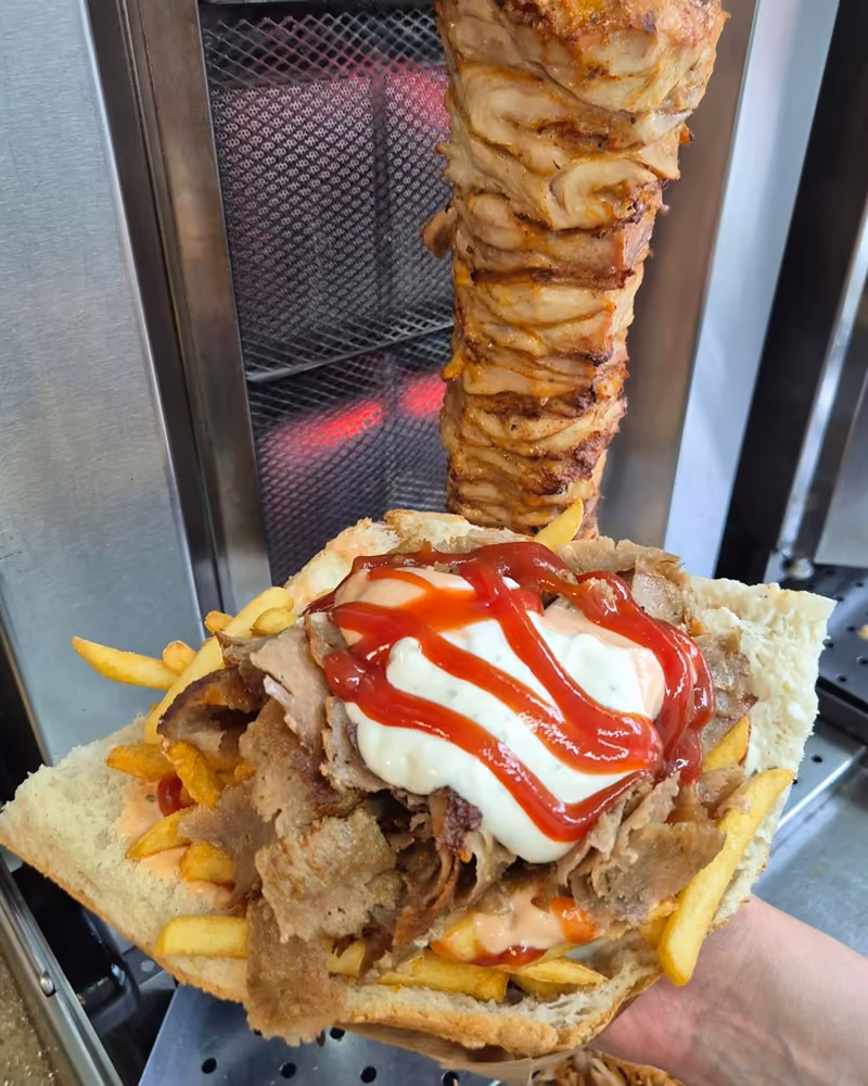 Kebab w bułce z frytkami i sosami trzymany na tle obracającego się rożna